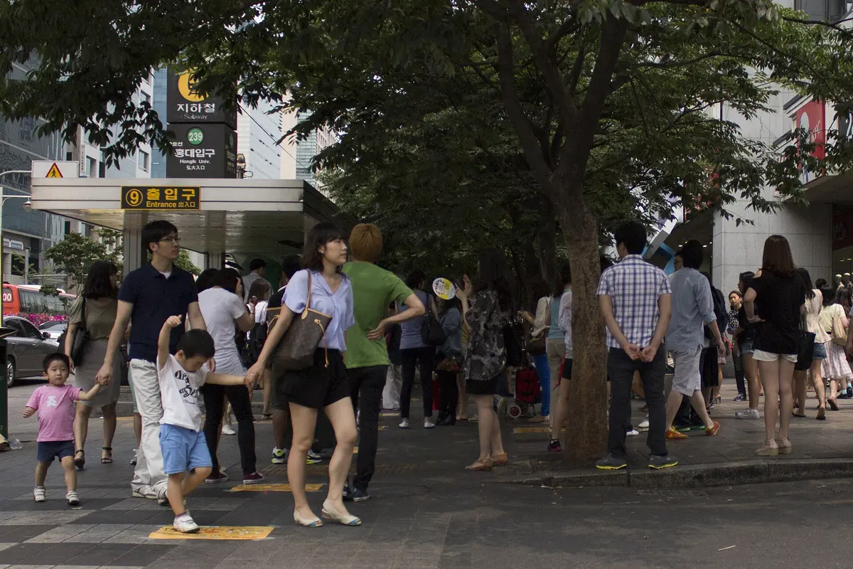 Exit 9 of Hongik University Station in Seoul, a covered subway stairwell with a crowd of people on the pavement and trees overhead