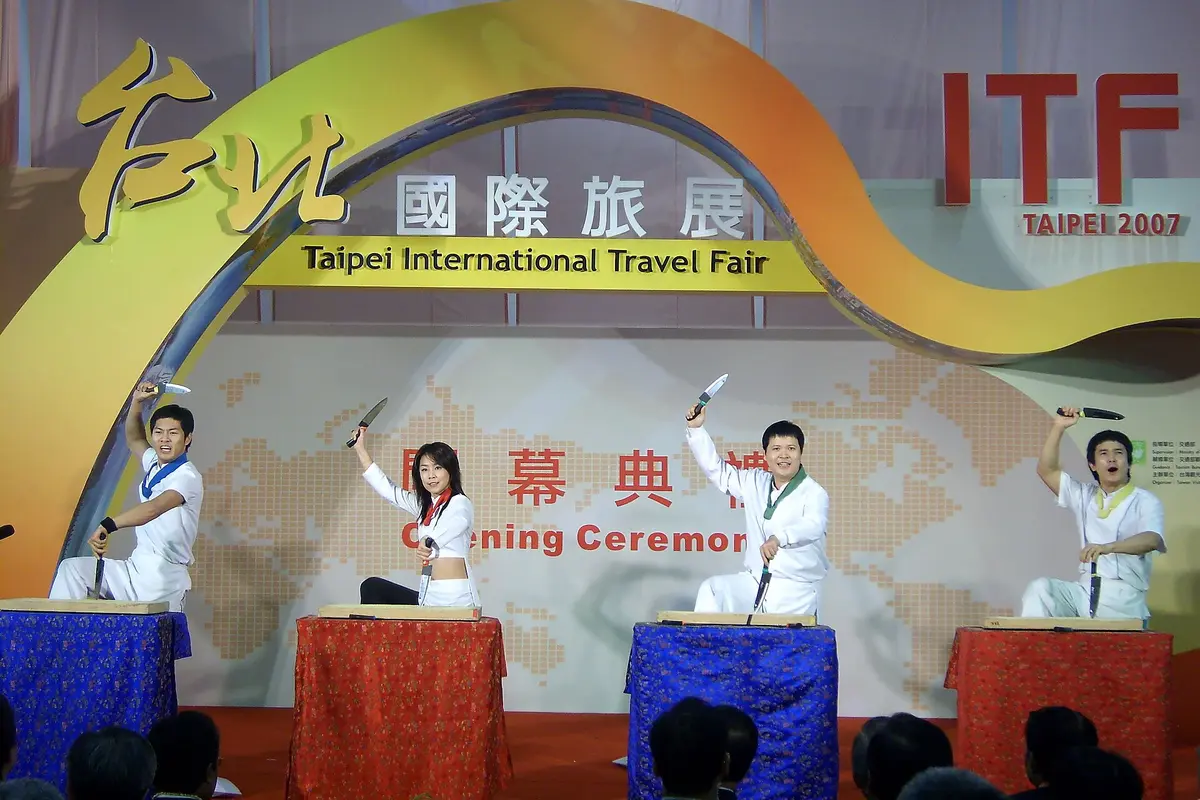 Four performers in white kitchen uniforms kneeling at tables, each raising a knife above a wooden cutting board on a stage