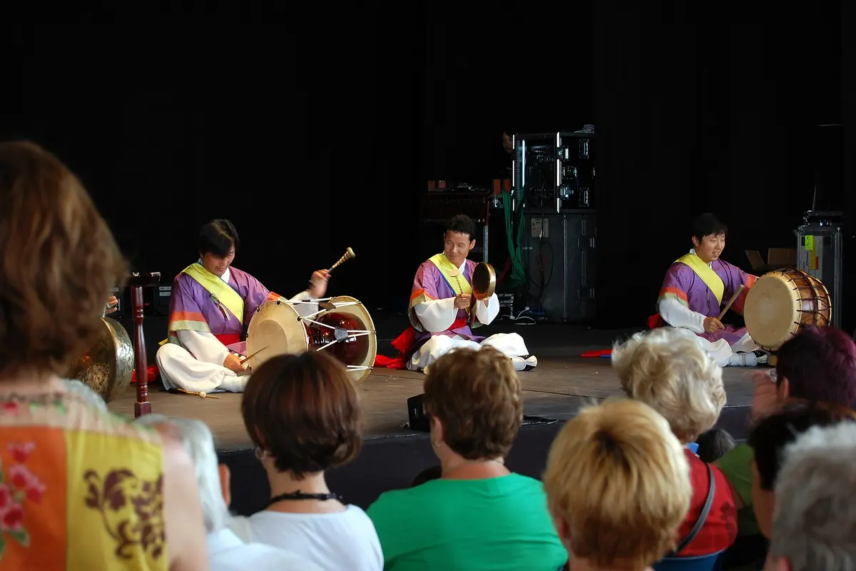 Three musicians in purple and white costumes sitting on a stage playing an hourglass drum, a small gong and a barrel drum, with an audience in the foreground