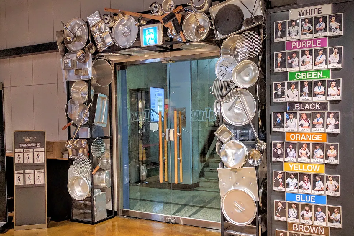Glass doors framed by steel pots, lids and trays, with a board of performer photos under coloured headings to the right